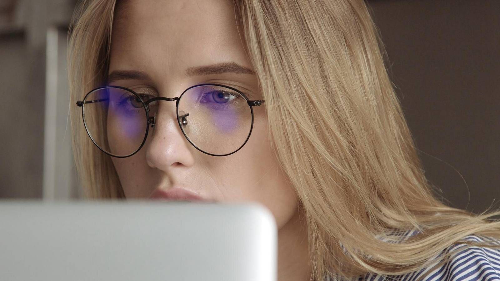 Close-up of a young woman with blonde hair wearing round, dark-rimmed glasses, looking intently at a laptop screen
